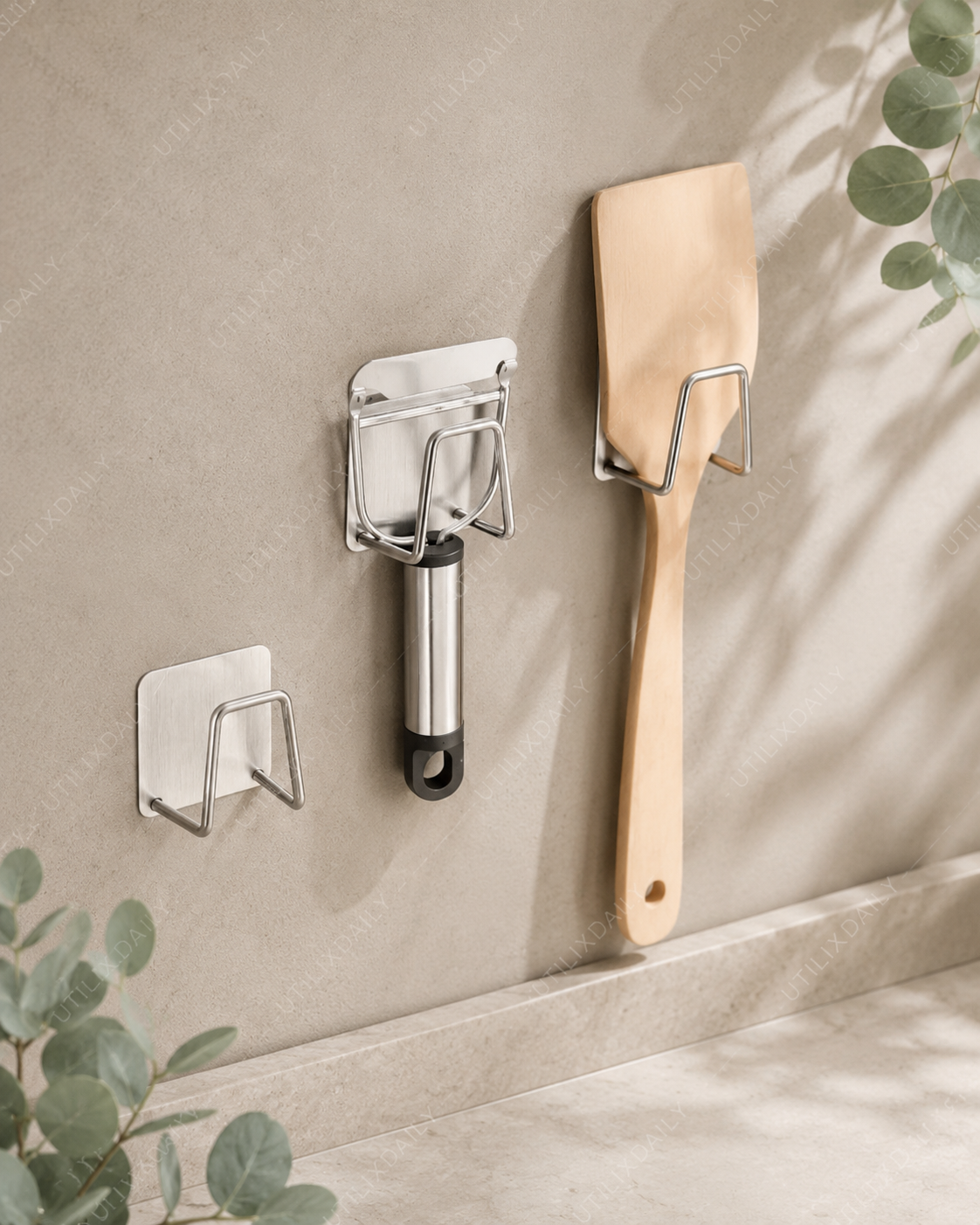 Silver self-adhesive kitchen hook holding a wooden utensil on a neutral wall with a clean minimalist style
